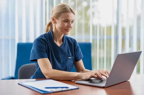 Nurse at desk working on a laptop with clipboard nearby