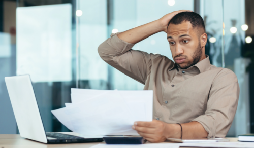 Man looking concerned while reviewing documents at a desk with a laptop in an office setting