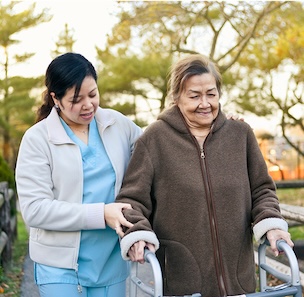 Caregiver assisting elderly woman with a walker outdoors in a park