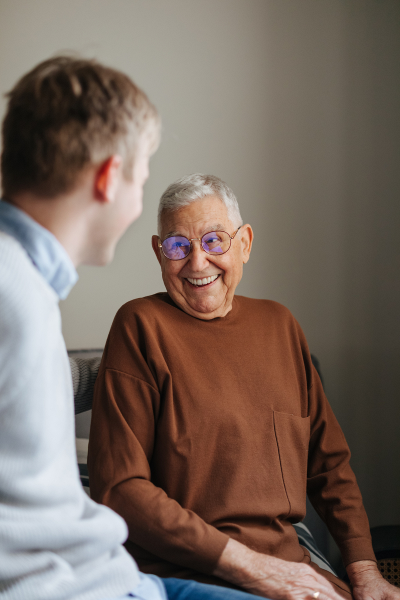 Older man smiling with glasses converses with a younger person indoors